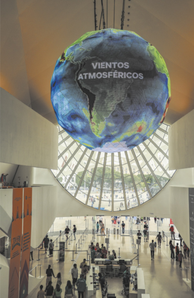 Fotografia do saguão de um museu, onde um globo terrestre com a inscrição 'Ventos Atmosféricos' está suspenso no teto, acima dos balcões de atendimento ao público. Diversas pessoas circulam pelo espaço.
