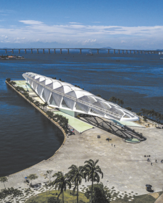 Fotografia aérea de um museu com arquitetura contemporânea, localizado sobre um píer que se estende sobre a água. A estrutura é alongada, com aparência futurista, formas assimétricas e cobertura em arco. Ao fundo, há uma extensa ponte sobre o mar e embarcações espalhadas pelas águas.