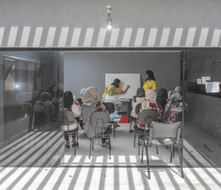 Fotografia de uma sala de aula vista de frente, com fachada de vidro e sombras lineares projetadas no chão. No interior, cadeiras estão organizadas em fileiras com várias pessoas sentadas, a maioria usando véus islâmicos na cabeça. Duas pessoas estão em pé diante de um quadro branco, uma escrevendo e a outra observando.