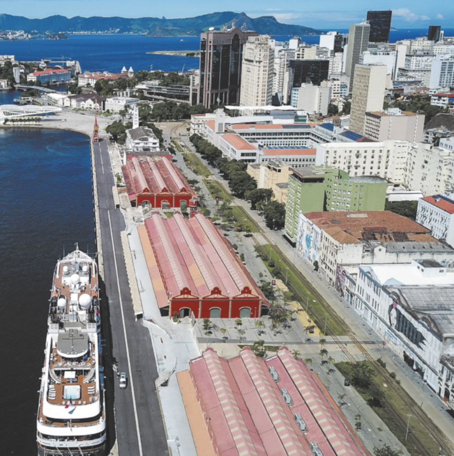 Fotografia aérea de uma área urbana à beira-mar, com galpões alinhados em fileira paralela à orla. Ao lado, encontra-se atracado um grande navio. Atrás dos galpões, observam-se edifícios de variadas alturas e trechos arborizados.