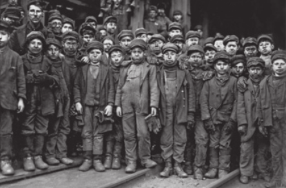 Fotografia em preto e branco de um grande grupo de meninos, todos com aparência jovem, reunidos em frente à entrada de uma mina. Eles vestem roupas de trabalho sujas e desgastadas, além de gorros e boinas. Muitos apresentam expressões sérias e cansadas. Um homem adulto posa ao lado dos meninos para a foto.