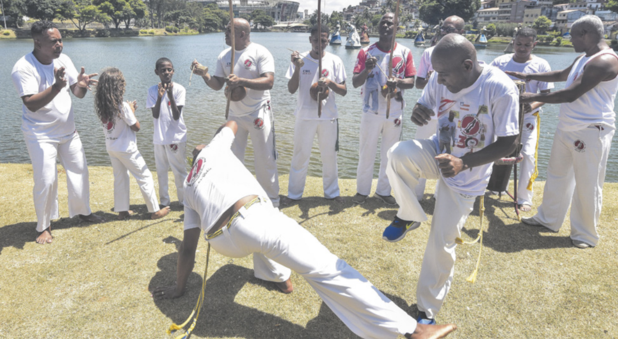 Fotografia de uma roda de capoeira em um parque, à beira de um lago. As pessoas vestem roupas brancas, tocam berimbaus e batem palmas, enquanto dois homens praticam capoeira.