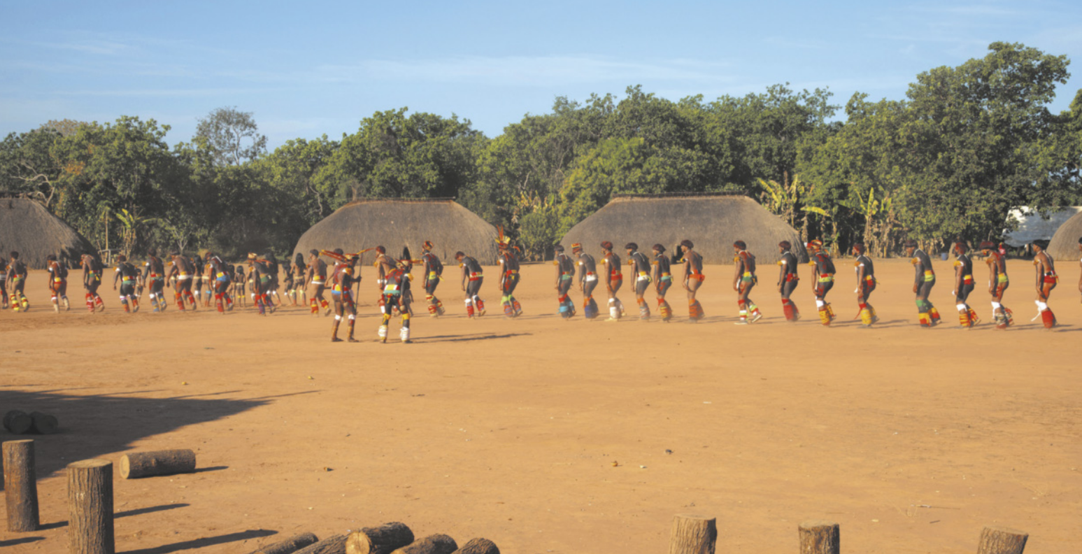 Fotografia de um grupo de pessoas indígenas enfileiradas em um espaço amplo de terra batida, realizando uma dança tradicional. Estão com os corpos pintados, vestindo trajes típicos e cocares. Ao fundo, há construções cobertas por palha e uma área de mata densa.