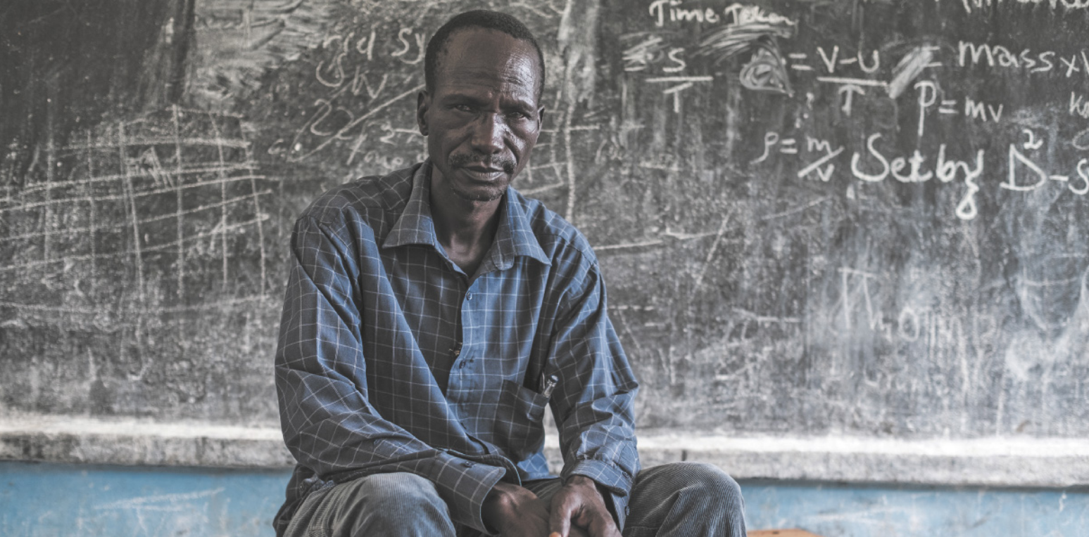 Fotografia de um homem negro com expressão séria, vestindo camisa de manga comprida e calça, sentado sobre uma mesa, tendo ao fundo uma lousa com marcas de giz.