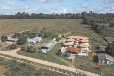 Fotografia aérea de uma área rural com diversas construções distribuídas ao longo de uma estrada de terra. As edificações estão cercadas por campos abertos e áreas de vegetação, com mata densa ao fundo.
