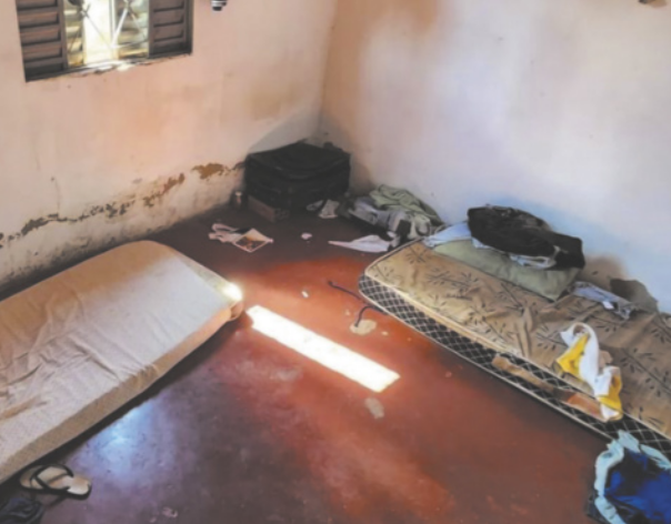 Fotografia de um cômodo com dois colchões em mau estado dispostos diretamente no chão. Há algumas peças de roupa espalhadas próximas a uma mala. As paredes apresentam marcas de mofo e o piso está inacabado, ainda no contrapiso.