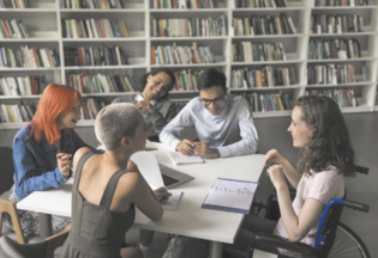 Fotografia de um grupo de estudantes sentados ao redor de uma mesa em uma biblioteca, conversando, sorrindo e escrevendo em seus cadernos.