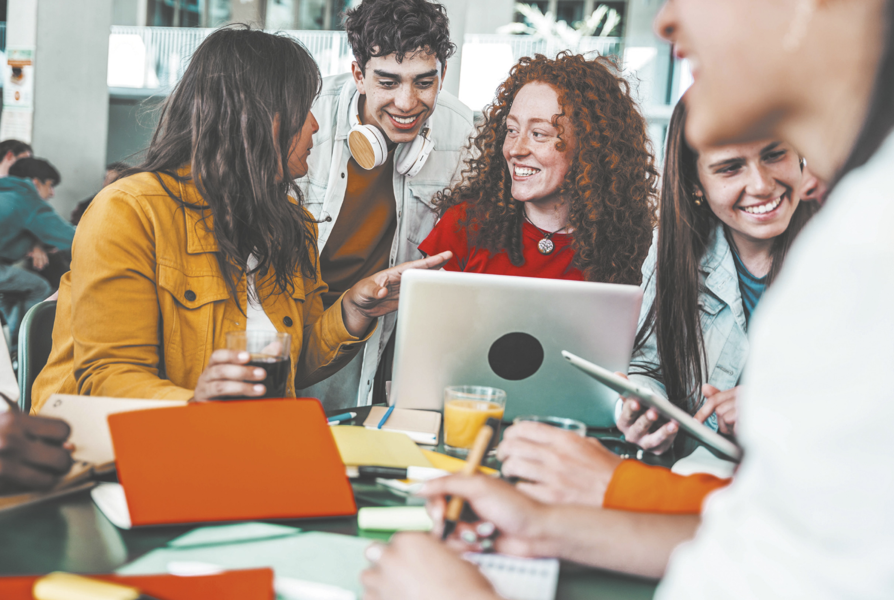 Fotografia de um grupo de jovens sentados ao redor de uma mesa, sorrindo e conversando enquanto interagem com um notebook e materiais de estudo.