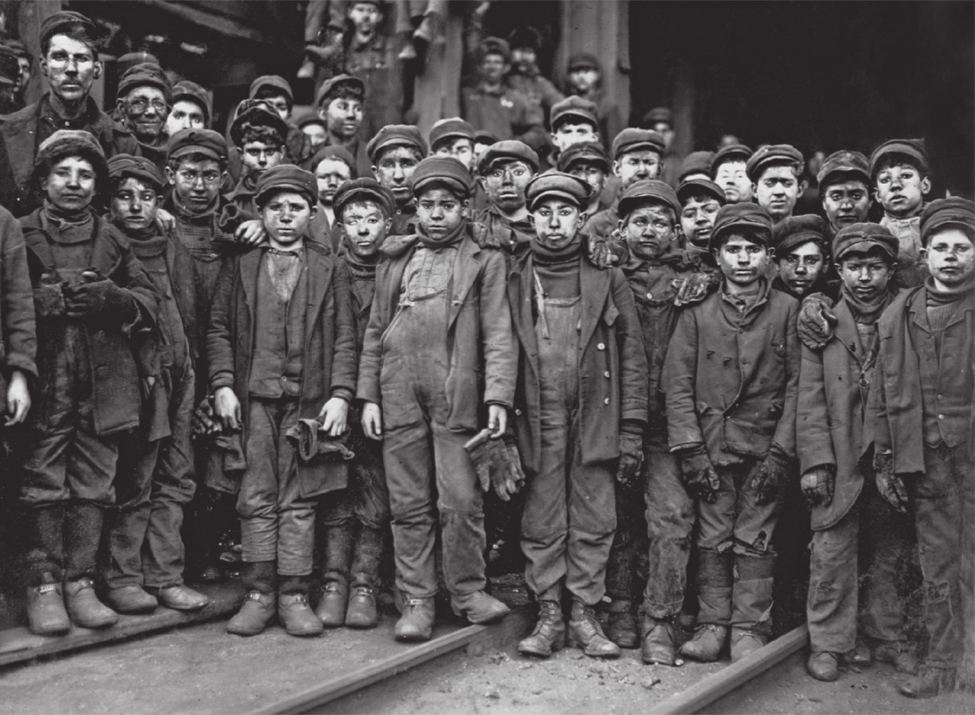 Fotografia em preto e branco de um grande grupo de meninos, todos com aparência jovem, reunidos em frente à entrada de uma mina. Eles vestem roupas de trabalho sujas e desgastadas, além de gorros e boinas. Muitos apresentam expressões sérias e cansadas. Um homem adulto posa ao lado dos meninos para a foto.