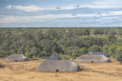 Fotografia aérea de uma área com ocas indígenas cobertas por palha, dispostas sobre um terreno amplo e plano. Ao fundo, há uma extensa área de mata fechada