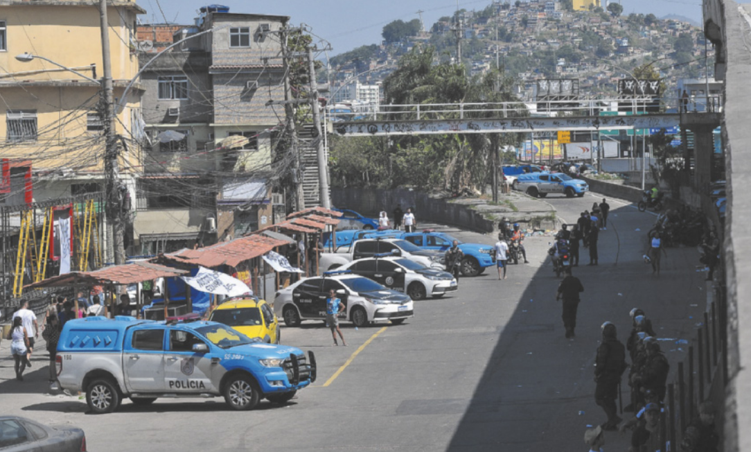 Fotografia de uma rua ao lado de uma comunidade com construções improvisadas e comércio popular, com barracas distribuídas pela calçada. Diversas viaturas estão estacionadas na rua e algumas pessoas circulam pelo local.
