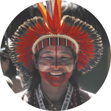 Fotografia de uma mulher indígena sorrindo, usando um cocar de penas na cabeça e com pinturas corporais no rosto.