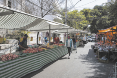Fotografia de uma feira livre, com barracas de frutas e legumes e pessoas circulando com máscaras de proteção.