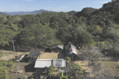 Fotografia aérea de uma pequena área rural cercada por mata, com algumas construções simples espalhadas pelo terreno. Ao fundo, é possível ver uma cadeia de montanhas.