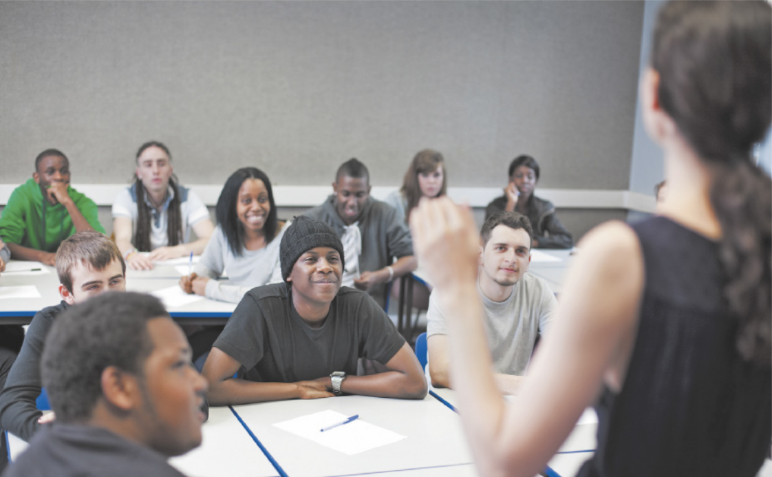 Fotografia de uma sala de aula com estudantes sentados em carteiras, voltados para uma pessoa em pé à frente, que está falando. Os estudantes demonstram atenção e alguns sorriem.