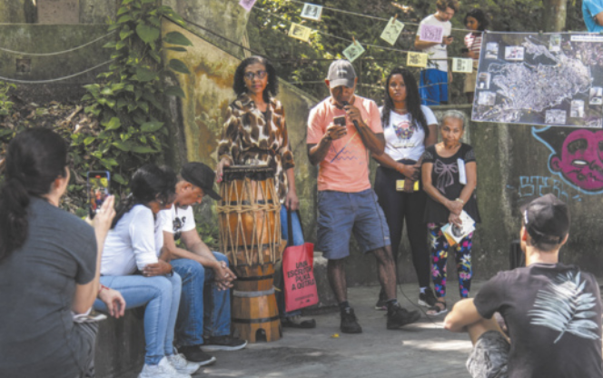 Fotografia de um grupo de pessoas reunido em um espaço ao ar livre, cercado por vegetação e muros decorados com cartazes e folhetos de cordel pendurados em barbantes suspensos. Algumas pessoas estão sentadas, enquanto outras permanecem em pé, próximas a um atabaque colocado no chão. À frente, um homem próximo ao atabaque fala ao microfone enquanto lê algo no celular.