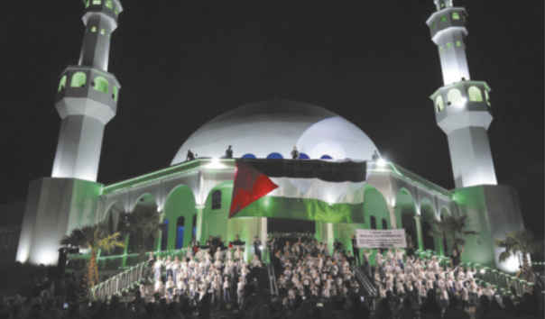 Fotografia de uma mesquita com duas torres simétricas e a bandeira da Palestina projetada em sua fachada, com várias pessoas reunidas na escadaria à frente.