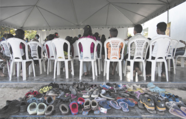 Fotografia de um grupo de pessoas sentadas em cadeiras plásticas sob uma tenda. Elas estão de costas, voltadas para uma atividade à frente. No chão, à frente da tenda, há muitos pares de calçados infantis e adultos organizados em fileiras.