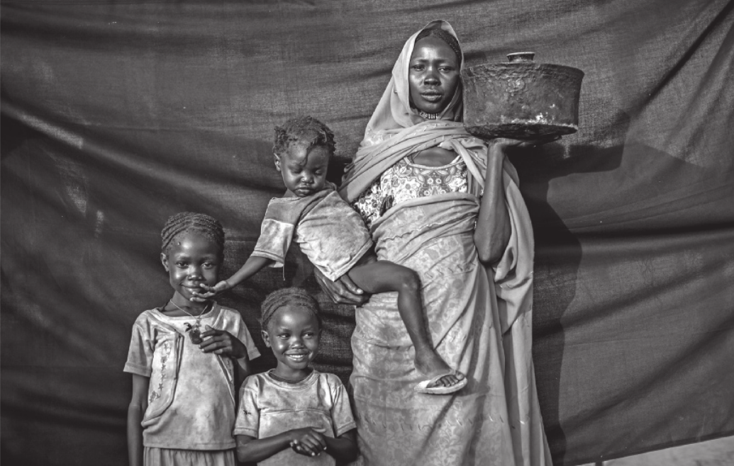 Fotografia de uma mulher negra posando com três crianças negras. Ela está com o corpo coberto por tecidos e usa um véu islâmico que cobre os cabelos. Segura uma panela em uma das mãos e carrega uma das crianças no outro braço. As outras duas estão paradas ao seu lado, sorrindo.