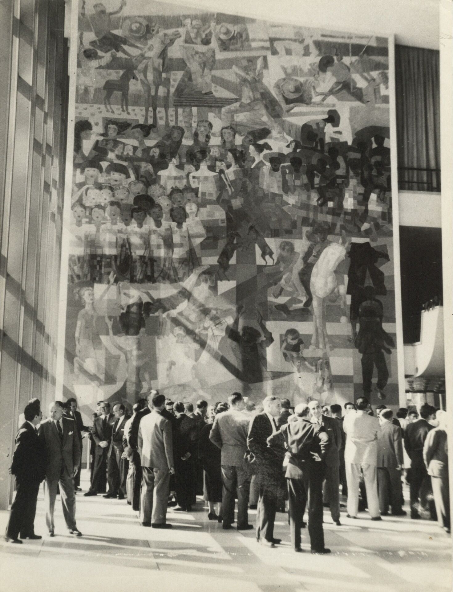 Fotografia em preto e branco. Em primeiro plano, diversos homens de costas, todos vestem terno. Em segundo plano, um grande painel composto por pinturas de pessoas e animais sobrepostas.