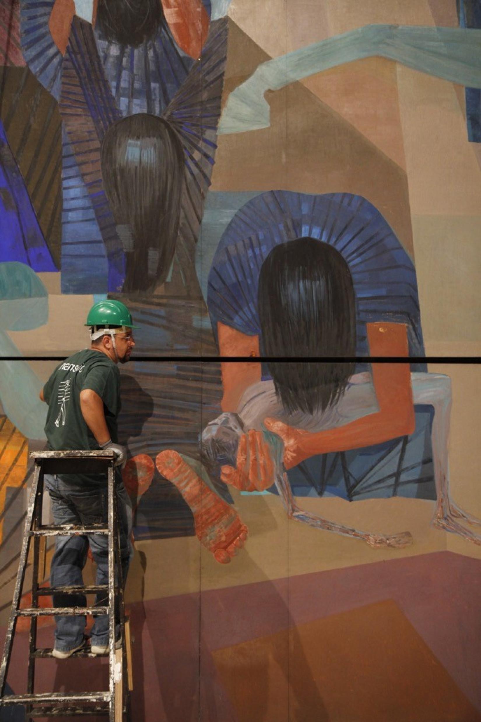 Fotografia. À esquerda, um homem de capacete verde, camiseta preta, calça jeans e tênis está de costas, em cima de uma escada. Na sua frente, um grande painel com a pintura de uma pessoa de roupa azul e cabelo preto, inclinada para frente com o cabelo cobrindo o rosto. Ela segura o corpo de uma criança nua.