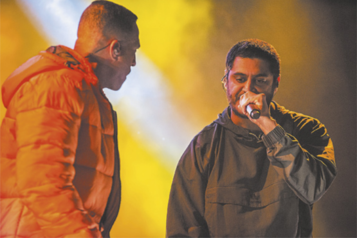 Fotografia de Mano Brown e Criolo em uma apresentação. Criolo é um homem de cabelos curtos e canta ao microfone. Mano Brown é um homem de cabelo raspado, usa uma jaqueta laranja e está ao lado de Criolo.