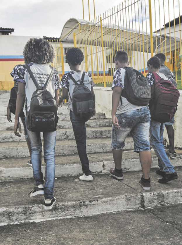 Fotografia de cinco estudantes de costas, carregando mochilas nas costas, subindo uma escada em direção à entrada de uma escola.