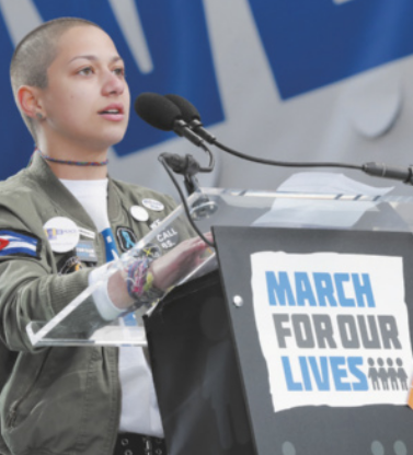 Fotografia de Emma González. Ela é uma mulher branca, de cabelos raspados, usa uma jaqueta com muitos broches e está falando em um púlpito. No púlpito, há um cartaz, escrito em inglês 'Marcha pelas nossas vidas'.