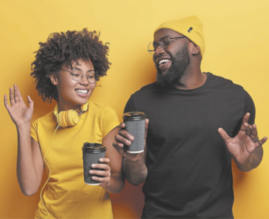 Imagem de dois jovens, um ao lado do outro, em pé, com uma parede amarela ao fundo. Do lado esquerdo, temos uma menina negra de cabelos curtos e encaracolados, usando uma blusa amarela e um fone de ouvido amarelo no pescoço. Ela está com olhos e boca fechados, sorrindo, segurando um copo térmico na altura da cintura. Sua mão direita está erguida e ela olha para o lado. Ao seu lado, há um homem negro, usando uma touca amarela e uma blusa preta. Ele está com os olhos fechados e boca fechados, segurando um copo térmico com a mão direita.