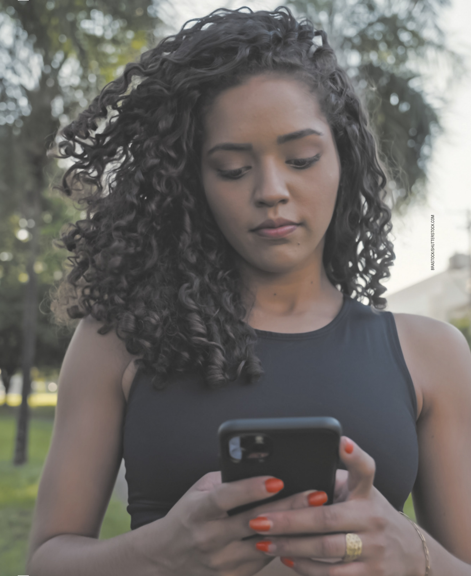 Fotografia de uma jovem negra de cabelos longos e cacheados, em pé em um parque, usando o aparelho celular.