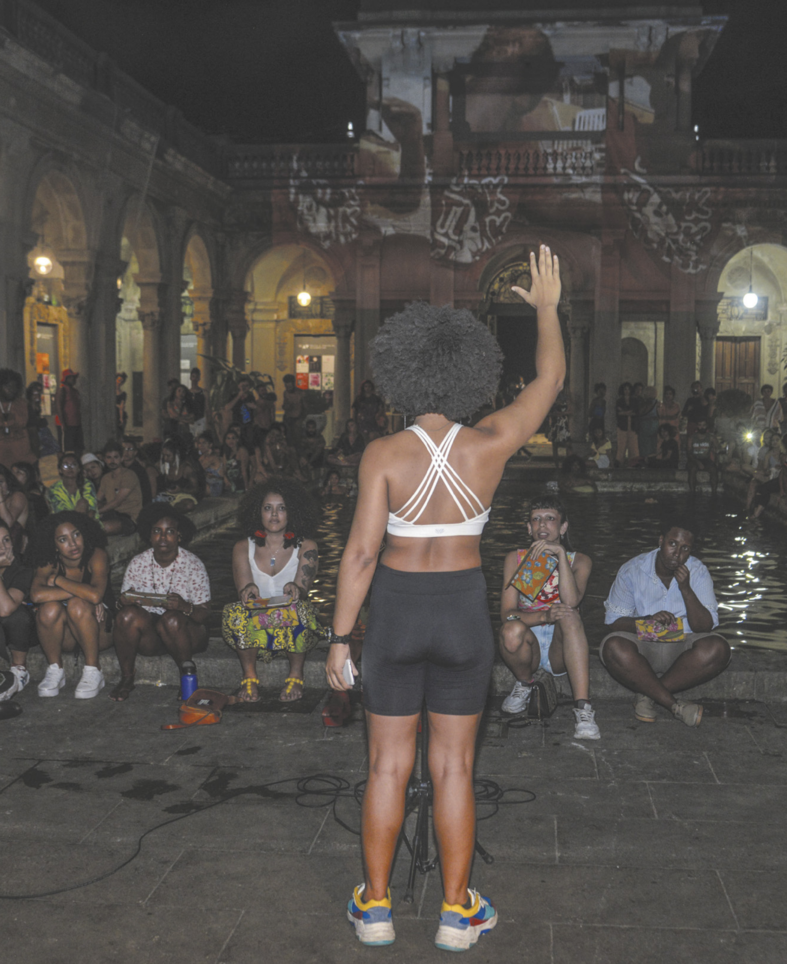 Fotografia de uma apresentação em área aberta, à noite. Uma menina negra está em pé, de costas, com o braço estendido. À sua frente, há um grupo de pessoas sentadas, assistindo.