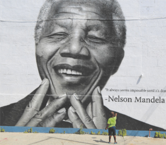 Fotografia do rosto de Nelson Mandela pintado em um muro, em preto e branco. Nelson Mandela é um homem negro, de cabelos curtos e grisalhos e está com os olhos fechados, levando as mãos ao rosto e sorrindo.