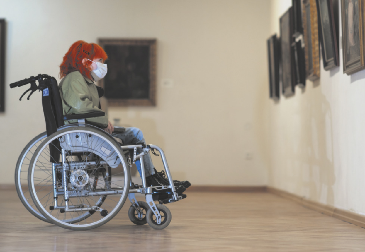 Fotografia de uma jovem de cabelos longos e vermelhos, sentada em uma cadeira de rodas, observando quadros expostos nas paredes de um museu. A menina usa uma máscara de proteção cobrindo boca e nariz.
