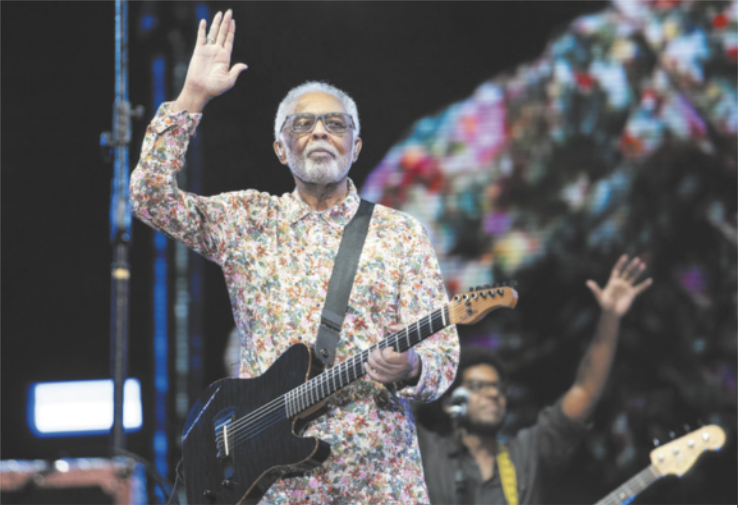Fotografia de Gilberto Gil. Ele é um homem negro, de cabelos e barba grisalhos e está se apresentando em um show, segurando uma guitarra e acenando com as mãos.