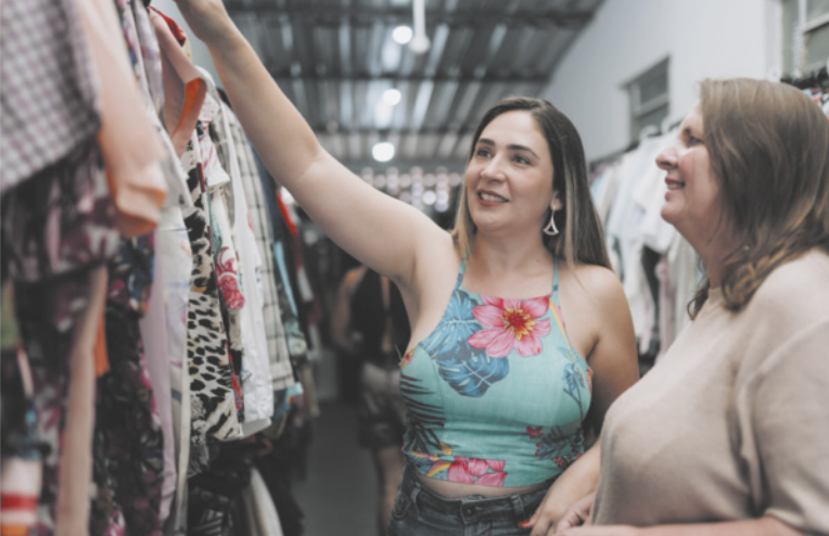 Fotografia de duas mulheres em uma loja observando roupas expostas em araras.