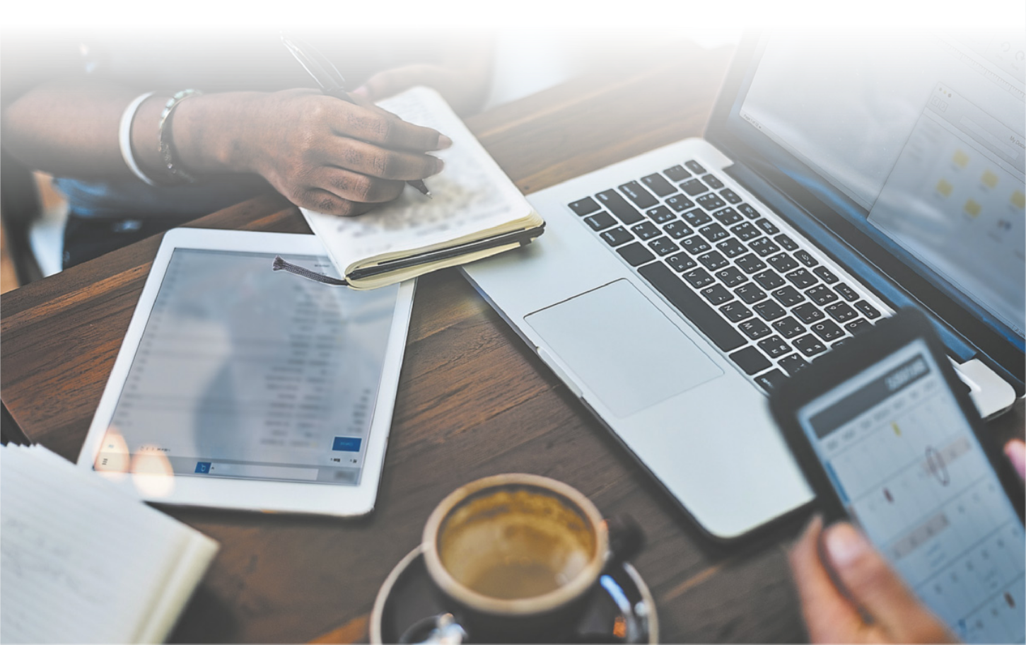 Fotografia de uma mesa com um notebook aberto, uma xícara de café e pessoas usando aparelhos celulares e tablets.