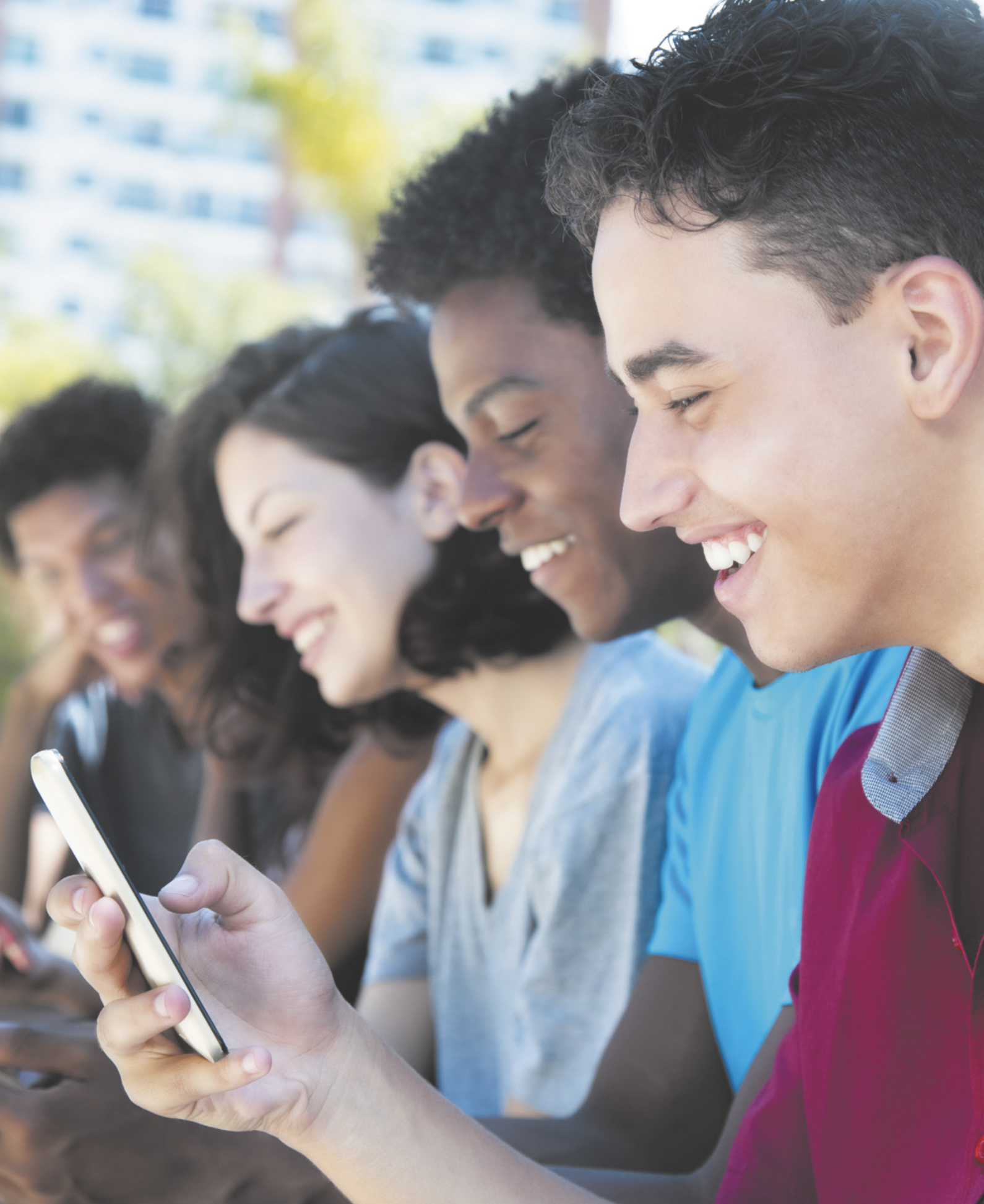 Fotografia do rosto de quatro jovens sentados lado a lado, cada um interagindo com seu próprio celular e sorrindo.