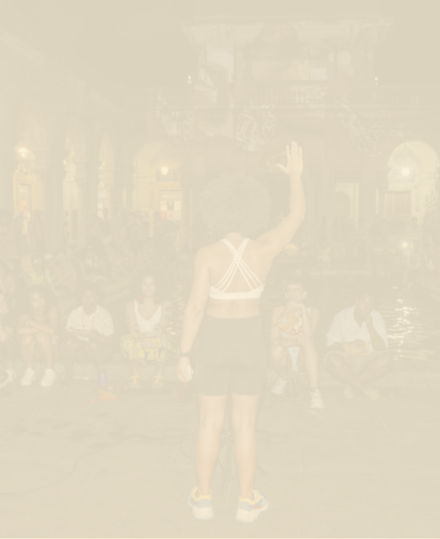 Fotografia desfocada de uma apresentação em área aberta, à noite. Uma menina negra está em pé, de costas, com o braço estendido. À sua frente, há um grupo de pessoas sentadas, assistindo.