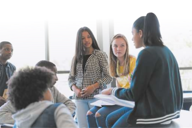 Fotografia de um grupo de jovens, meninos e meninas, reunidos em uma sala, conversando. Uma menina dialoga gesticulando com as mãos, enquanto os outros a observam atentamente.