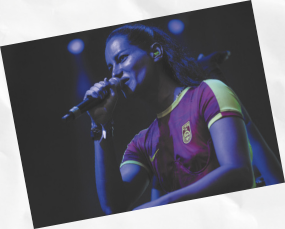 Fotografia de Jaloo. Ela é uma mulher branca, de cabelos compridos e está cantando em um microfone.