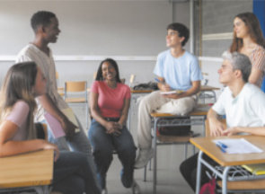 Fotografia de um grupo de jovens, homens e mulheres, reunidos em uma sala. Eles estão sentados em círculo e em carteiras, conversando.