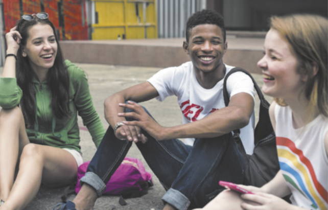 Fotografia de três jovens, duas meninas e um menino, sentados no chão, sorrindo e conversando de forma descontraída.