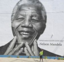 Fotografia do rosto de Nelson Mandela pintado em um muro, em preto e branco. Nelson Mandela é um homem negro, de cabelos curtos e grisalhos e está com os olhos fechados, levando as mãos ao rosto e sorrindo.
