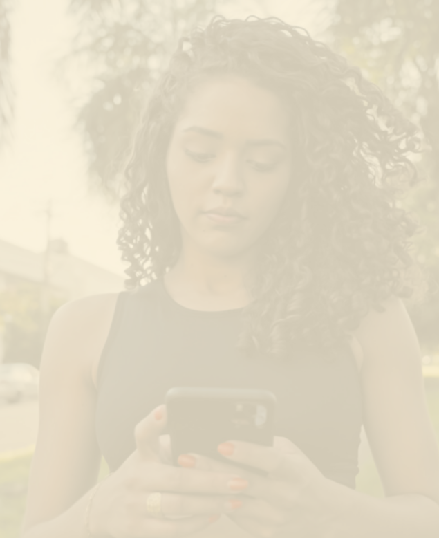 Fotografia desfocada de uma jovem negra de cabelos longos e cacheados, em pé em um parque, usando o aparelho celular.