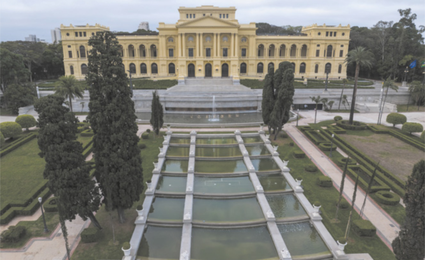 Fotografia da fachada do Museu do Ipiranga. Ele é um edifício longo, com formato retangular e simétrico, composto por três andares visíveis. A superfície do prédio é lisa e pintada em tom amarelo claro. Na parte inferior da fachada, há uma grande escadaria central que leva até a entrada principal. A entrada possui portas grandes e arqueadas, com janelas acima delas. Ao longo da fachada, existem várias colunas verticais distribuídas de forma regular. Entre as colunas, há janelas altas com forma arqueada na parte superior. No nível superior da fachada, há um elemento triangular no centro. Todo o edifício está cercado por jardins com caminhos e plantas alinhadas.