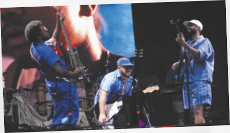 Fotografia da banda Os Gilsons, formada por três homens negros, um tocando baixo e outros dois tocando guitarra. Eles se apresentam em um show, em cima de um palco.