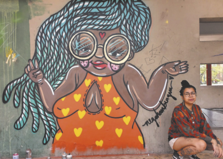 Fotografia de Evelyn Queiróz. Ela é uma jovem negra, com os cabelos escuros, presos em um coque e está sentada no chão, ao lado de um grafite desenhado na parede. O grafite representa uma jovem negra de óculos, cabelos longos e trançados, usando um vestido laranja com corações amarelos.