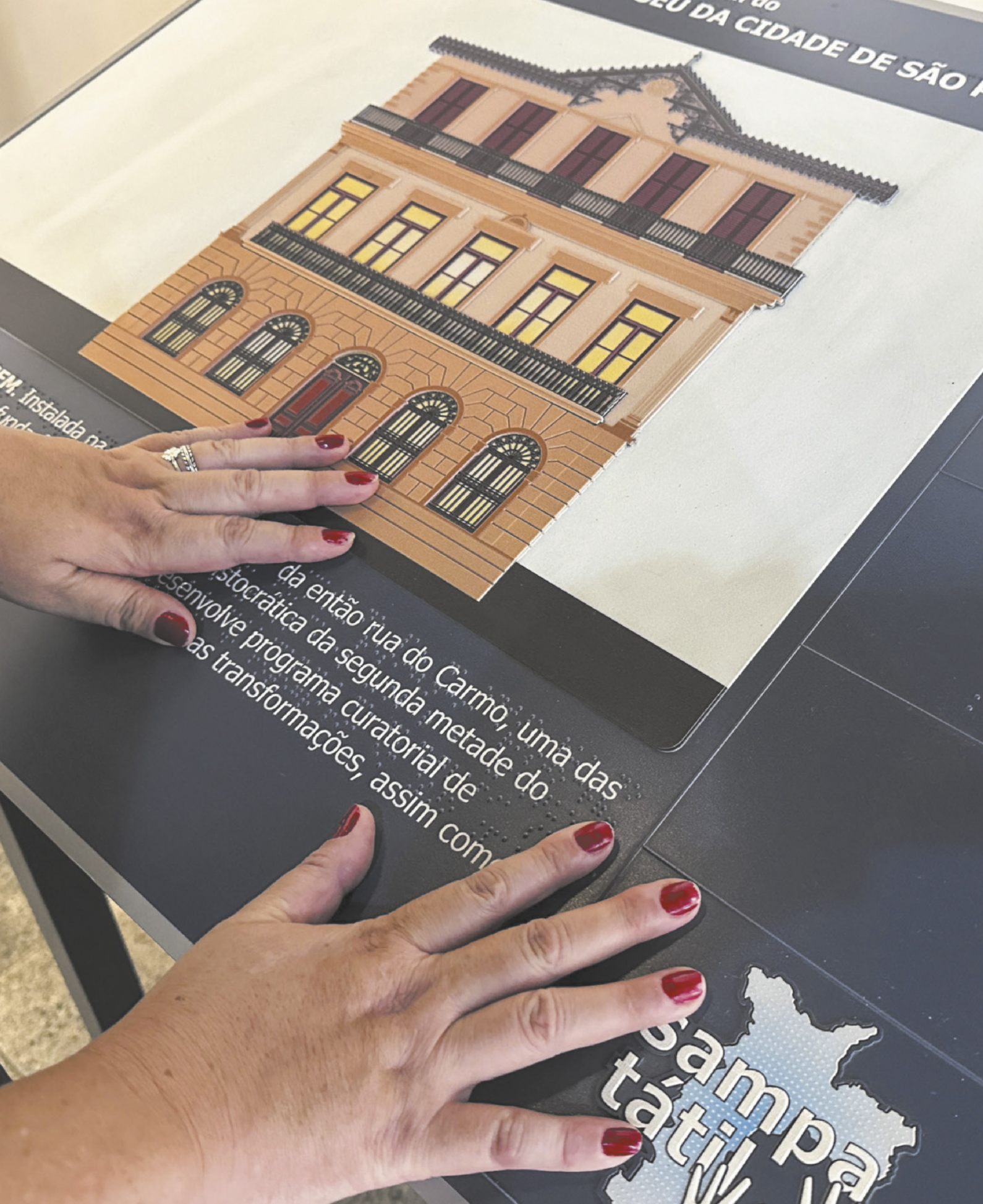 Fotografia de uma mulher usando as mãos para ler um mapa tátil com a imagem de um museu e um texto em Braille.