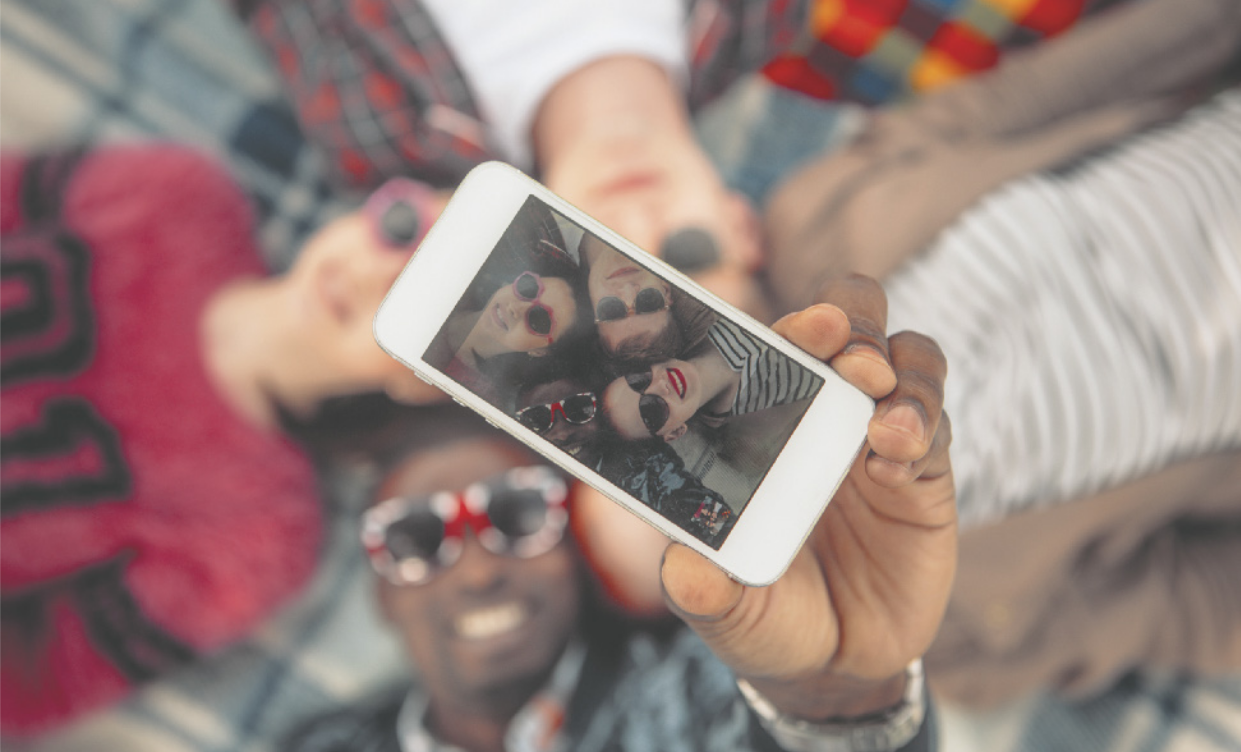 Imagem da tela de um celular com a fotografia do rosto de um grupo de quatro jovens deitados no chão, usando óculos escuros. Ao fundo, observa-se o grupo de jovens na mesma pose da foto.