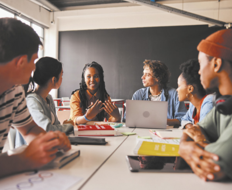 Fotografia de um grupo de jovens sentados em frente a uma mesa retangular, olhando uns para os outros, discutindo ideias. Uma menina fala, gesticulando com as mãos, enquanto o restante do grupo a observa. Um menino está com o notebook aberto e há livros e folhas em cima da mesa.
